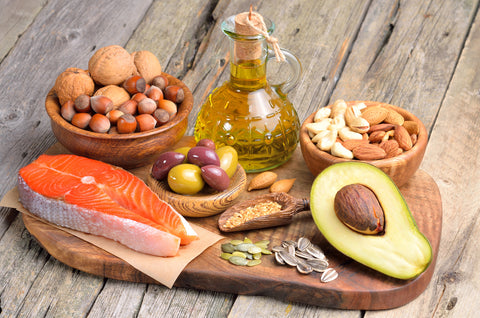 Ein Brett voller Lebensmitteln aus oder mit gesunden Fetten: Avocado, Lachs, Olivenöl, Haselnüsse, Walnüsse, Mandeln, Chashewkerne, Oliven und Samen