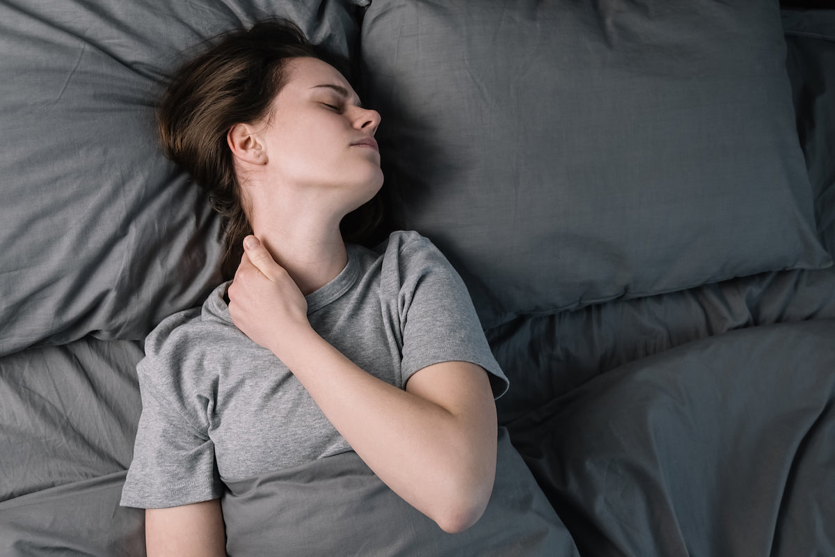 Frau liegt im Bett und greift sich vor Schmerzen an den Nacken