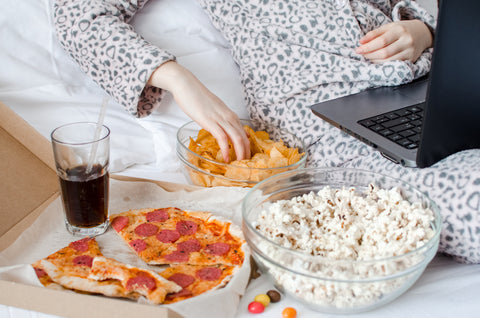 Jemand sitzt mit Laptop im Bett und isst dabei Chips und Pizza
